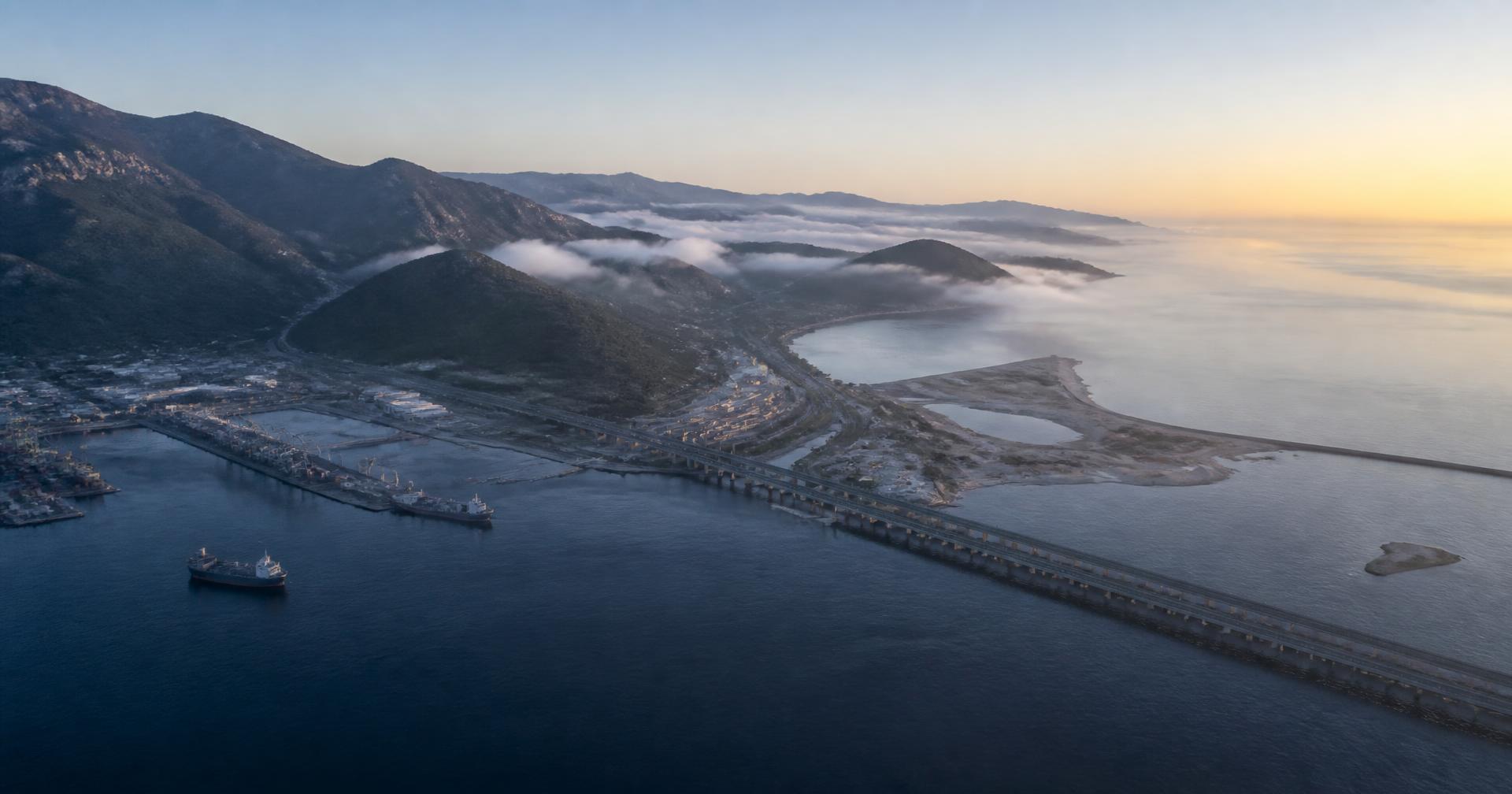 Aerial view at dawn of a coastline where a port, causeway and roads meet mountainous terrain